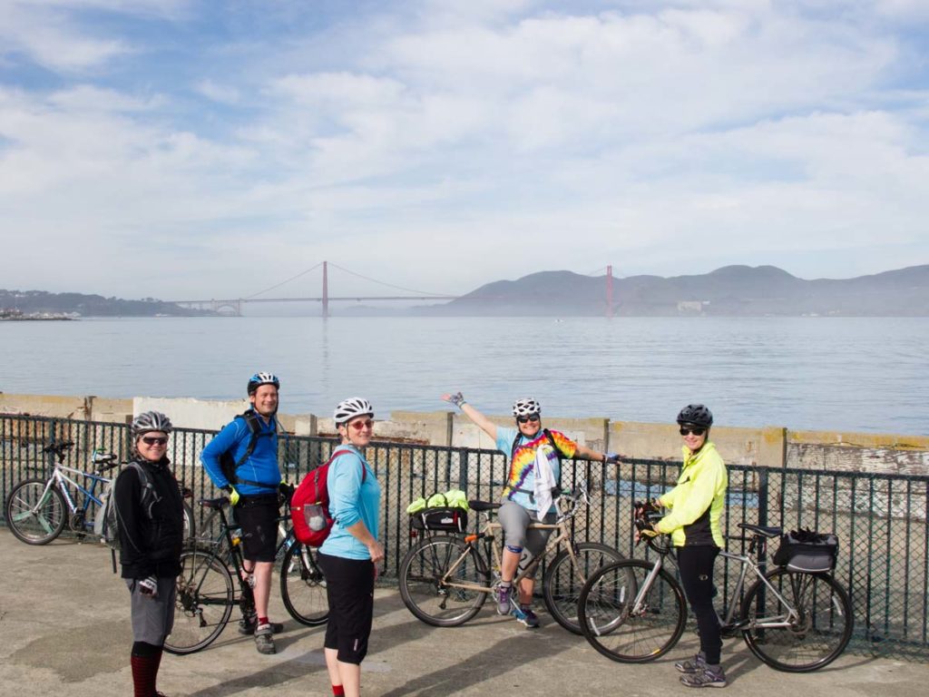 A group of cyclists stands at a seawall with the Golden Gate Bridge in the background on a misty morning. They are all smiling. One is pointing to the bridge.